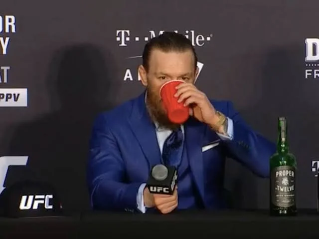 Conor McGregor drinking from red cup with Proper Twelve Irish whiskey bottle at UFC press conference